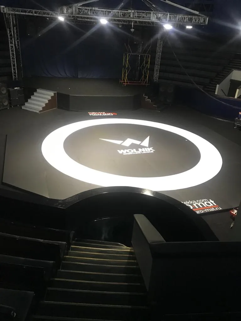 Серый борцовский ковер с логотипом WOLNIK и белой круговой разметкой на арене с трибунами