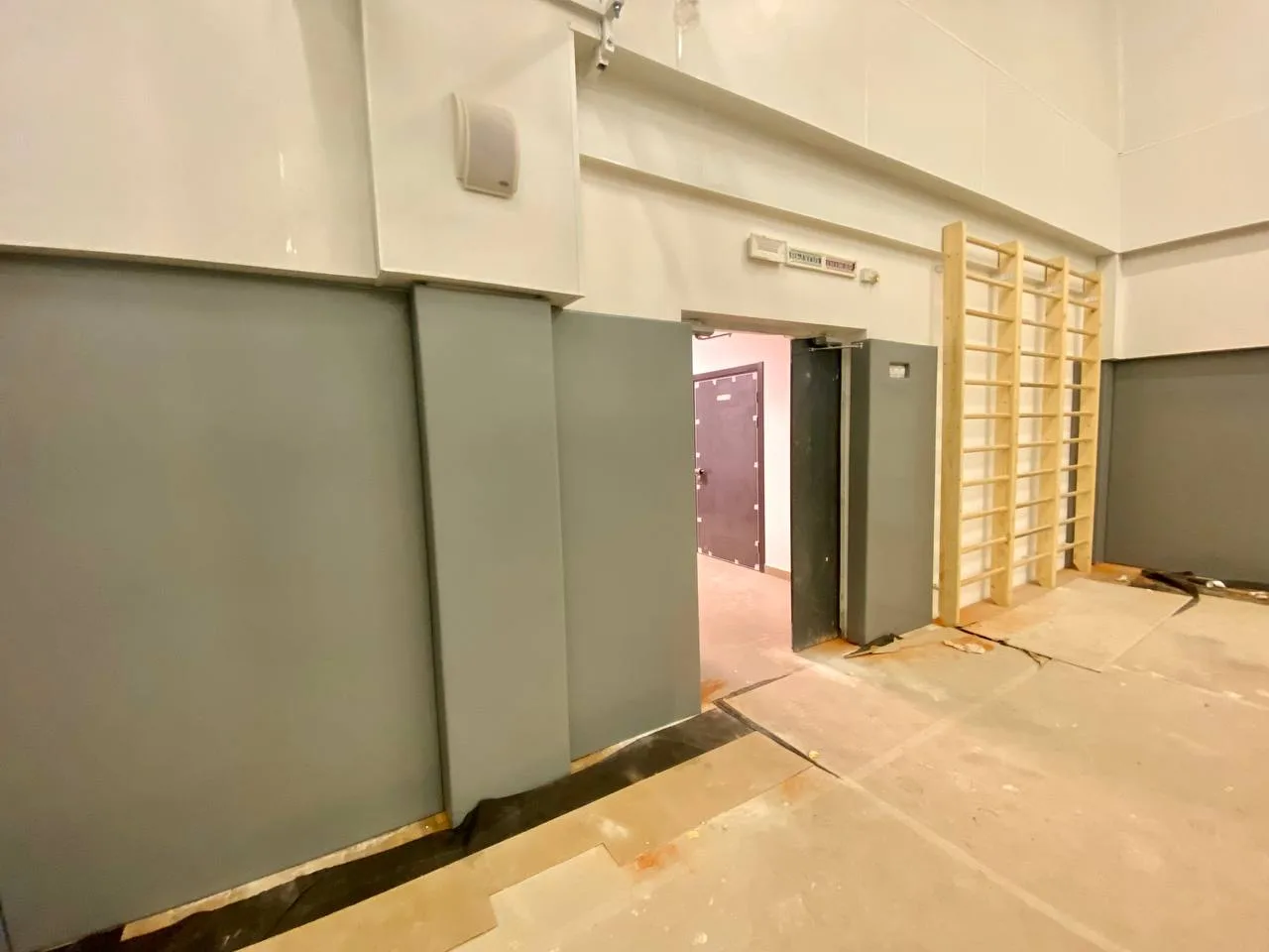 Серые стеновые протекторы у входной двери школьного спортзала, гимнастические лестницы на стене