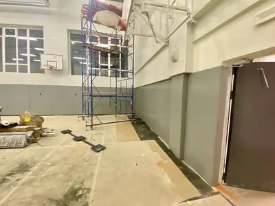 Угол школьного спортзала с серыми протекторами, строительными лесами и баскетбольным кольцом