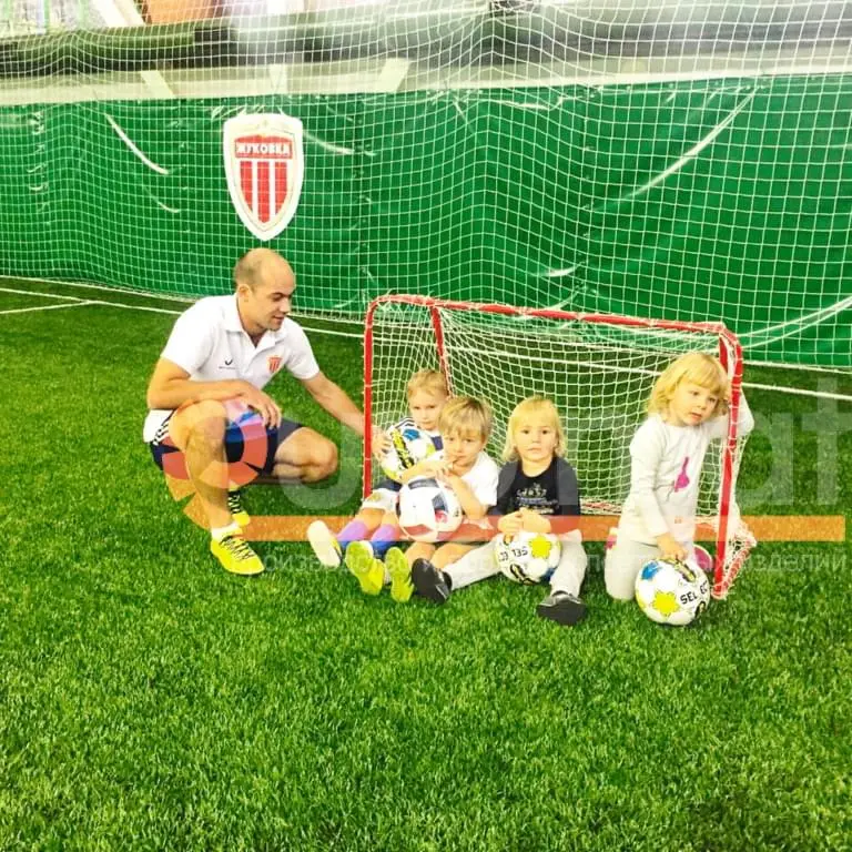 Тренер с детьми у мини-ворот на фоне зелёных стеновых протекторов Жуковка