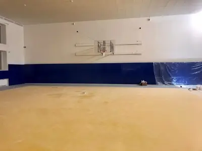 Синие протекторы под баскетбольным кольцом, жёлтый спортивный пол, частично закрытая стена справа