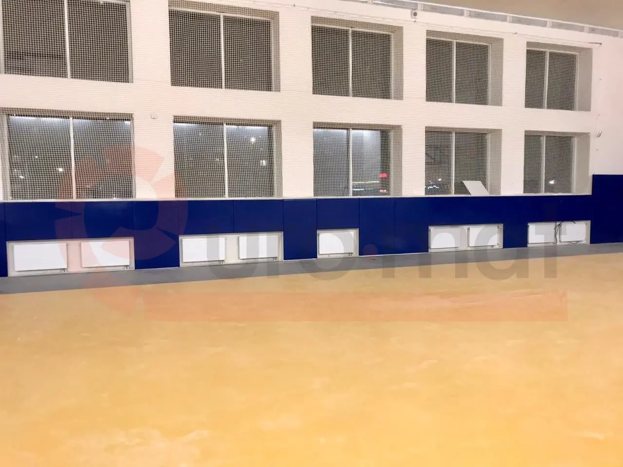 Синие стеновые протекторы вдоль стены зала единоборств с окнами и защитной сеткой, жёлтый пол