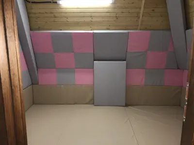 Розово-серые стёганые протекторы на стене детской комнаты с белыми матами на полу бревенчатого дома