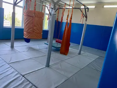 Угол зала адаптивной физкультуры с синими протекторами на стенах и серыми матами, вид на качели