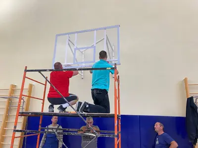 Установка баскетбольного щита на леса, рабочие вид сзади, синие протекторы на стенах школьного зала