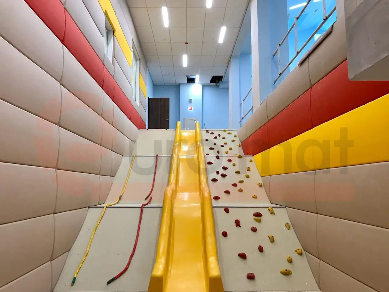 Бежевые стеновые протекторы по бокам скалодрома с жёлтой горкой в детском спортзале школы