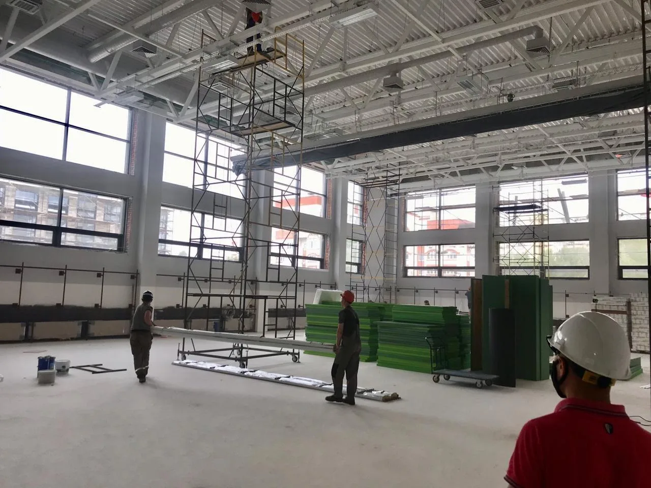 Большой школьный спортзал с зелёными протекторами в стопках и рабочими на лесах