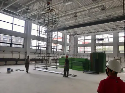 Большой школьный спортзал с зелёными протекторами в стопках и рабочими на лесах
