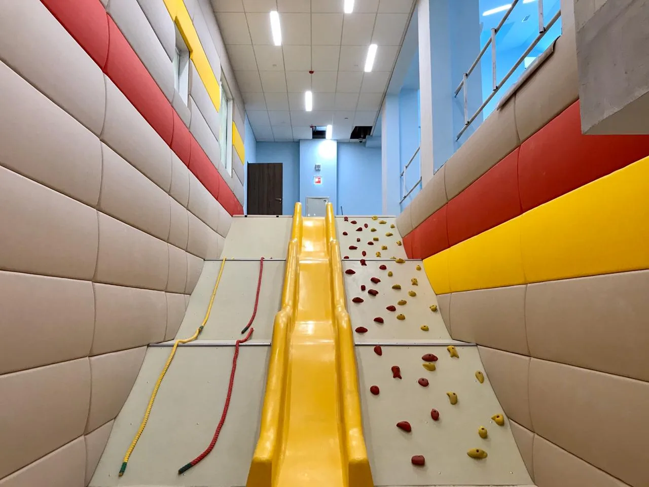 Жёлтая горка и скалодром между выпуклыми стеновыми протекторами в игровой зоне