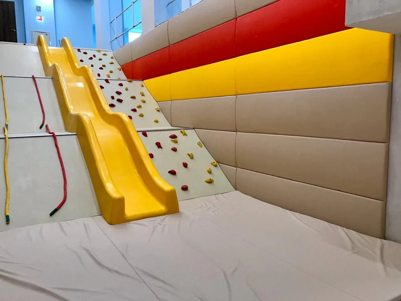 Жёлтая горка со скалодромом и выпуклые бежево-красно-жёлтые протекторы на стенах снизу
