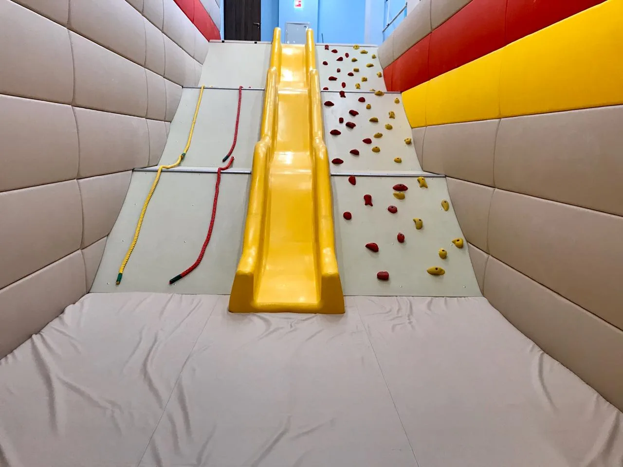 Горка и скалодром с мягким матом внизу, обрамлённые выпуклыми бежевыми протекторами